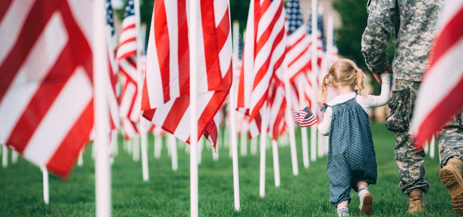 little girl walking with her dad in uniform among graves marked with American flags on Memorial Day